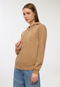 Felpa con zip color beige, dotata di tasca frontale, cappuccio con coulisse e polsini a coste. Indossata con jeans azzurri chiari e logorati. Texture liscia.