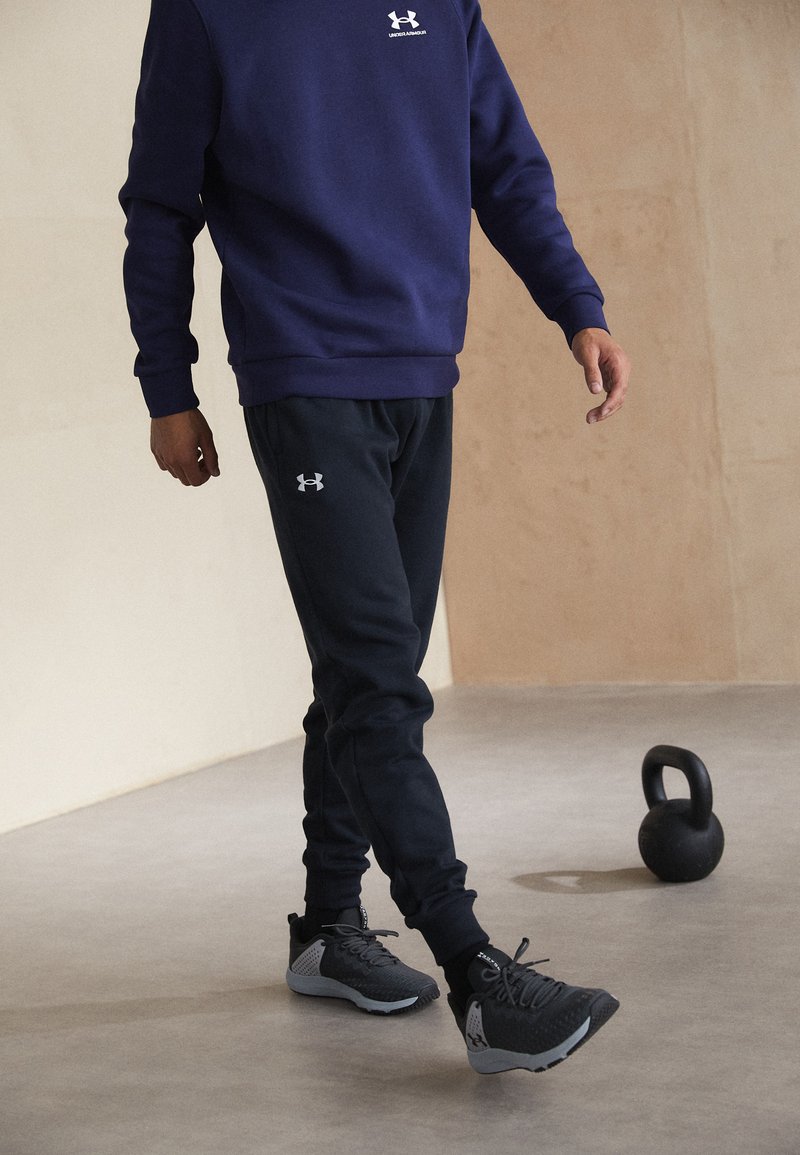 Marinblå sweatshirt och svarta joggers i mjukt material med vita logotyper. Svarta träningsskor med grå detaljer. Kettlebell bredvid modellen.