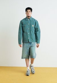 Veste en denim vert avec fermeture à boutons et poches poitrine, associée à un short en denim bleu clair. Le modèle porte des baskets grises avec des chaussettes blanches.