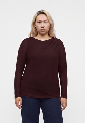 Femme aux longs cheveux blonds portant un haut à manches longues foncé et un jean sombre, debout devant un fond blanc uni.