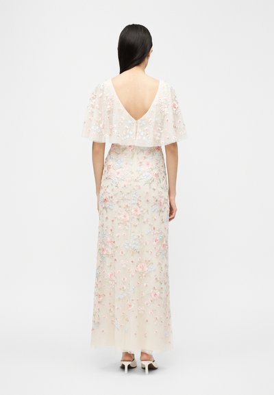 Γυναίκα με μακριά σκούρα μαλλιά που φοράει φόρεμα μήκους μέχρι το πάτωμα, κρεμ χρώματος με κεντήματα ροζ και μπλε ανθικών μοτίβων, με ακάλυπτη την πλάτη της, στέκεται μπροστά σε λευκό φόντο.