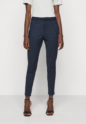 Personne portant un pantalon bleu marine slim, un haut à manches courtes rayé, et des sandales à talons noires avec bride à la cheville, debout devant un fond uni.