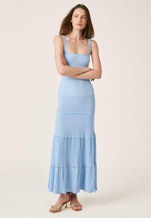 Mujer de pie con los brazos cruzados, vestida con un vestido maxi sin mangas de punto acanalado azul claro y sandalias de tacón alto beige.