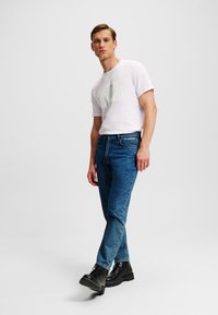 Biele grafické tričko s horizontálnymi pruhmi, kombinované s modrými džínsami. Model má na sebe čierne šnurovacie topánky. Neutrálne pozadie.