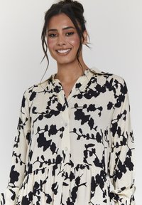 Abito camicia a maniche lunghe in crema con stampa botanica nera. Presenta un colletto, chiusura anteriore con bottoni e gonna plissettata per un tocco di texture in più.