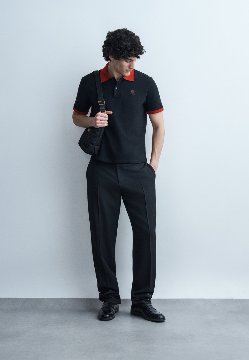 Young man in black trousers, black polo with red collar and cuffs, holding black shoulder bag, looking down against plain wall.