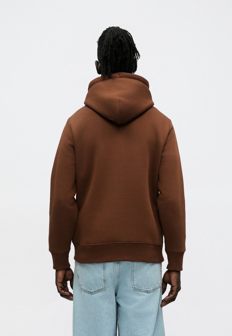 Brown hoodie made of soft fabric, featuring a large hood, ribbed cuffs, and hem. Worn with light blue denim jeans.