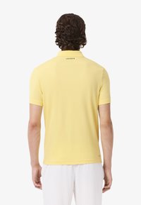 Polo gialla con colletto classico, maniche corte e piccolo logo Lacoste ricamato sulla schiena. Tessuto liscio e leggero.