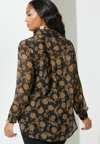 Musta läpinäkyvä paita kultaisella kukkakuviolla, pitkät hihat, kaulus ja napitettu hihansuu. Takana on pleat, joka lisää liikkuvuutta.