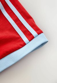 Czerwone szorty z niebieskimi ściągaczami, z dwiema niebieskimi paskami po bokach, wykonane z gładkiego bawełnianego materiału.