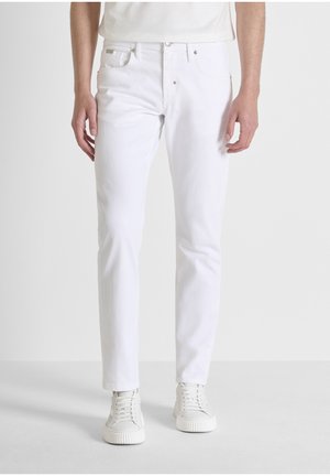 Hombre de pie con vaqueros blancos de corte ajustado y zapatillas blancas, sobre un fondo liso y claro.