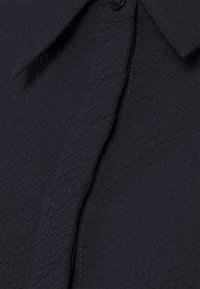 Gros plan d'un tissu texturé bleu marine foncé avec des motifs verticaux subtils et un col partiellement visible ainsi qu'une couture.