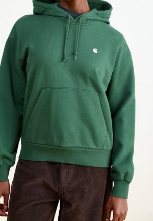 Osoba ubrana w zieloną bluzę z kapturem, z kieszenią na przodzie i małym białym logo, w połączeniu z ciemnobrązowymi spodniami sztruksowymi.