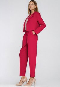 Fuksjowa krótka marynarka z długimi rękawami, w zestawie z dopasowanymi zwężanymi spodniami. Modelka ma na sobie cieliste szpilki, a pod marynarką widoczna jest fakturowana bluzka.