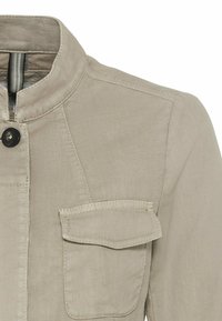 Beige jakke med opretstående krave, enkelt sort knaplukning og klaplomme på brystet.