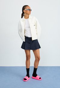 White zip-up jacket, white T-shirt, navy pleated skirt, black socks, pink slide sandals. Sunglasses with a patterned frame.