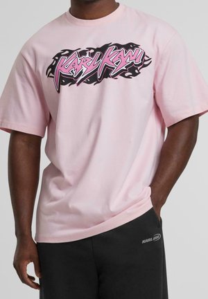 Man in een lichtroze T-shirt met zwart en roze "Karl Kani" graphic en zwarte broek met klein "Karl Kani" logo bij de zak.