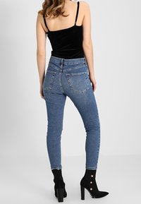 Džínové kalhoty světle modré barvy, s vysokým pasem, úzkého střihu s kapsami na zadní straně. V kombinaci s černým sametovým topem a kotníkovými botami se zlatými knoflíky.