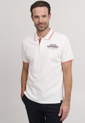 Camisa polo blanca con acentos rojos y detalle del logo en el lado izquierdo del pecho, combinada con pantalones oscuros. Fabricada en algodón suave, con mangas cortas.
