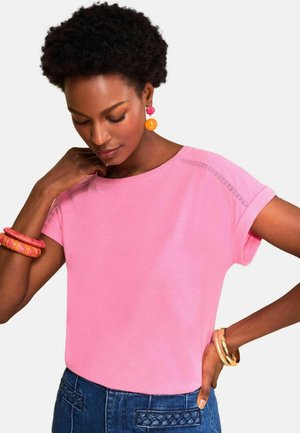 Lühikeste lokkis juustega naine, kes kannab roosat särki, siniseid džinse, värvilisi kõrvarõngaid ja käevõrusid, poseerides ühe käega puusal.