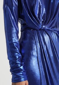 Lesklé modré šaty z látky s dlouhými, přiléhavými rukávy; mají záhyby a detail s otočením v pase. Hladká textura a kovový lesk.