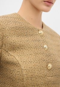 Guld- och gräddfärgad vävd tyg nära bild som har en rund halsringning och tre dekorativa knappar i guld. Texturen ser strukturerad och texturerad ut.