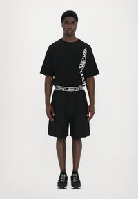 Svart kortermet t-skjorte med hvit "Versace Jeans Couture" logo, kombinert med svarte shorts og svarte joggesko med hvite detaljer.