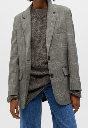 Person trägt graues kariertes Sakko über einem braunen Strickpullover und blauen Jeans vor einfachem Hintergrund.