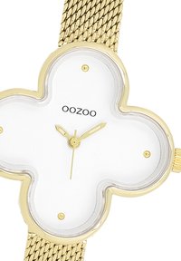Montre avec un bracelet en maille dorée et un boîtier en forme de trèfle. Cadran blanc et marqueurs d'heure dorés. Nom de la marque "OOZOO" affiché sur le cadran.