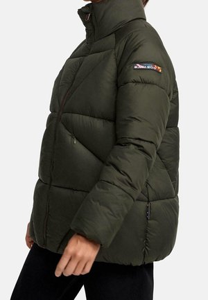 Personne portant une doudoune matelassée vert foncé avec un col montant et un logo en écusson sur la partie supérieure de la manche gauche.