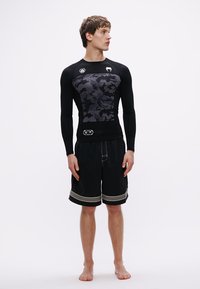 Czarna koszulka do pływania z długim rękawem, z szarym kamuflażowym wzorem i logo na piersi, w połączeniu z czarnymi kąpielówkami z żółtymi akcentami.