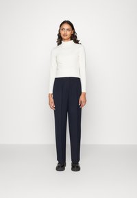 Top a collo alto fitted bianco a maniche lunghe abbinato a pantaloni plissettati blu scuro a vita alta. Scarpe nere con suola spessa completano l'outfit.