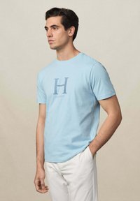 T-shirt en coton bleu clair à manches courtes, arborant une grande lettre bleue "H" et le logo "HACKETT" centré en dessous. Design simple à col rond.