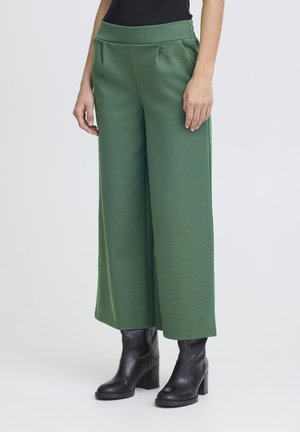 Mujer con pantalones anchos verdes combinados con botines negros de tacón, de pie contra un fondo liso de color gris claro.