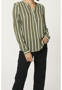 Personne portant une chemise à rayures verticales blanches et vert olive à manches longues avec des boutons, associée à un pantalon noir, se tenant avec une main dans la poche.