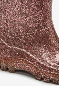 Gros plan de la partie inférieure d'une botte de pluie rose pailletée avec une semelle texturée et une surface brillante.