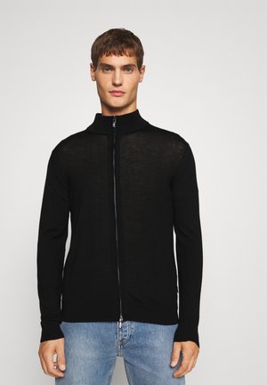 Cárdigan negro con cremallera, hecho de material ligero, presenta un cuello alto y un diseño ajustado, combinado con vaqueros azules.