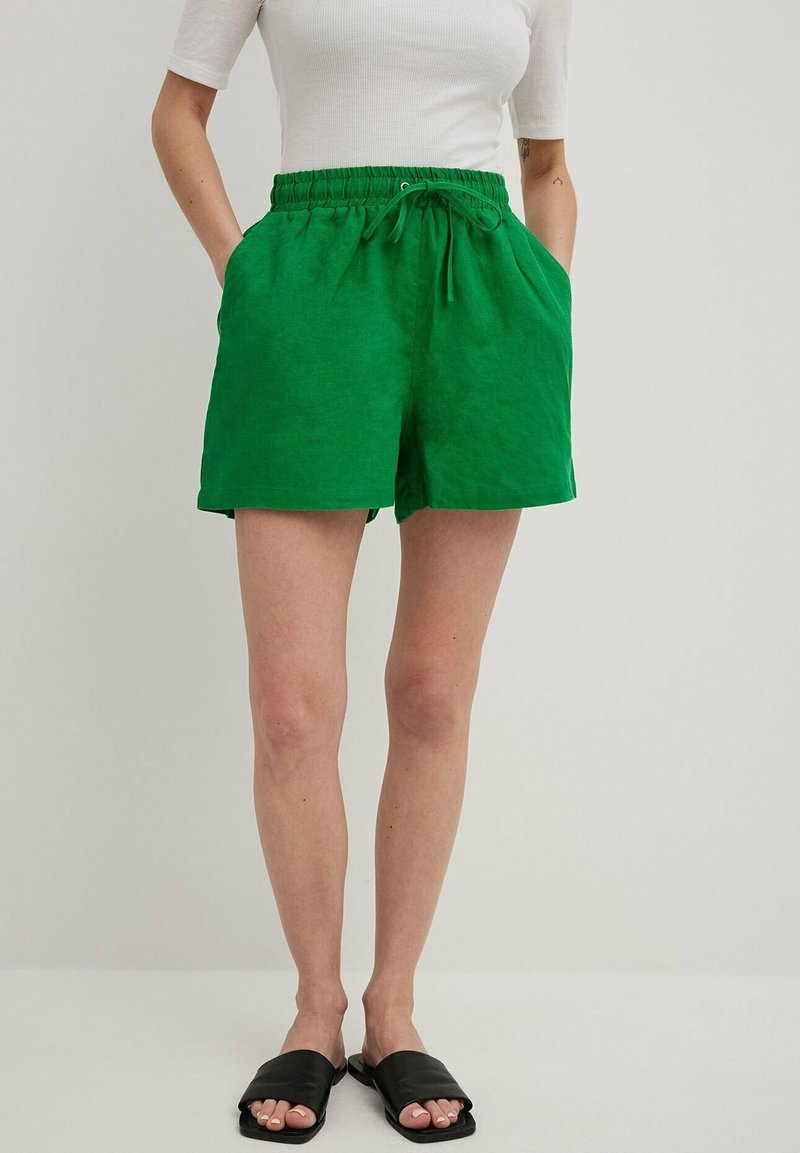 Groene linnen shorts met een elastische tailleband en trekkoord, voorzien van zijzakken en een relaxte pasvorm. G gedragen met zwarte instap sandaal.