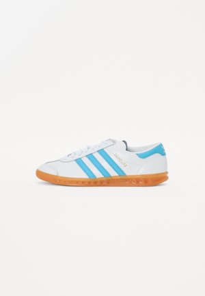 Zapatilla blanca con tres franjas azules, suela de goma naranja, parte superior de cuero suave, punta redondeada y logo en el costado y en el talón.