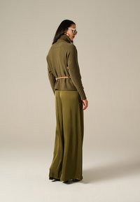 Maglione a collo alto in maglia verde oliva dalla forma a peplo, con cintura in vita, abbinato a pantaloni a gamba larga coordinati in un tessuto liscio.