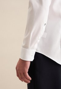 Witte textuurshirt met een aansluitend ontwerp. Kenmerkt zich door een knoopmanschet, subtiele borduursels en een gladde afwerking.