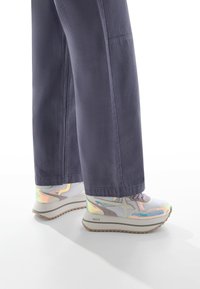 Sneakers bianchi e iridescenti con dettagli grigi e suola texture, abbinati a pantaloni scuri larghi, mostrando una vista laterale parziale.