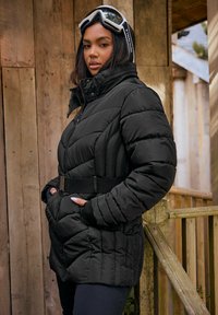 Schwarze Steppjacke mit hohem Kragen, tailliertem Bund, eng gesteppten Nähten und zwei vorderen Taschen. Getragen mit weißen Skibrillen.