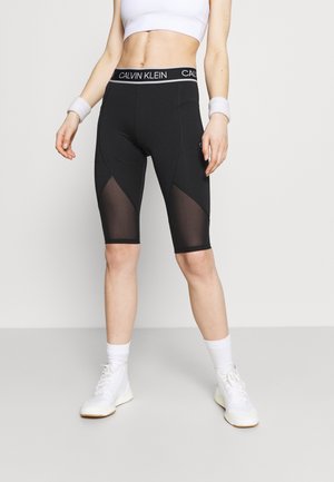 Kvinde iført sorte Calvin Klein biker shorts med meshpaneler, hvid crop top, hvide sokker, armbånd og hvide sneakers stående på en lys baggrund.