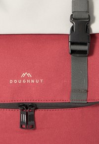 Rød og hvit stoffsekk med sydd logo "DOUGHNUT," med svarte glidelåsgrep og et grått justerbart belte med plastspenne.