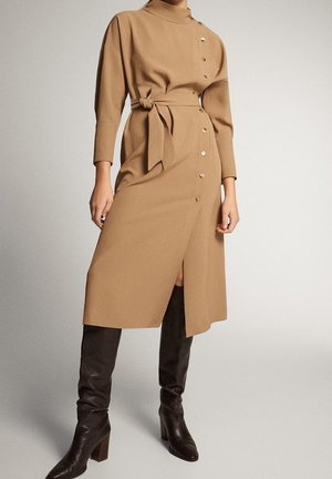 Femme portant une robe midi couleur chameau avec des boutons sur le côté, une ceinture à la taille et des manches longues, assortie à des bottes à talons noires montant jusqu'aux genoux.