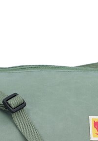 Saco de lona verde com uma superfície texturizada, apresentando uma alça com fivela preta e uma etiqueta de logotipo amarela e vermelha. Costura visível ao longo das bordas.