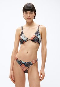 Kobieta o krótkich ciemnych włosach, ubrana w kolorowy bikini w geometryczne wzory, stojąca na tle jednolitego białego tła.