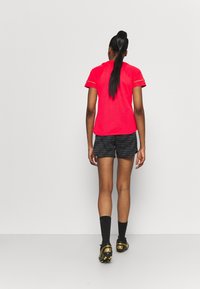 Röd atletisk tröja med korta ärmar, svarta mönstrade shorts, svarta strumpor och benskydd med gula detaljer. Sett bakifrån.