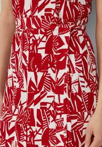 Rotes und weißes, abstrakt gemustertes Kleid mit einem Taillengürtel, abgebildet auf einer Person von der Brust bis zur Mitte des Oberschenkels, mit einem sichtbaren Arm.